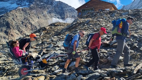 Eine Wandergruppe mit steigt mit einer Person in einem Treckingrollstuhl hinauf zu einer Berghütte